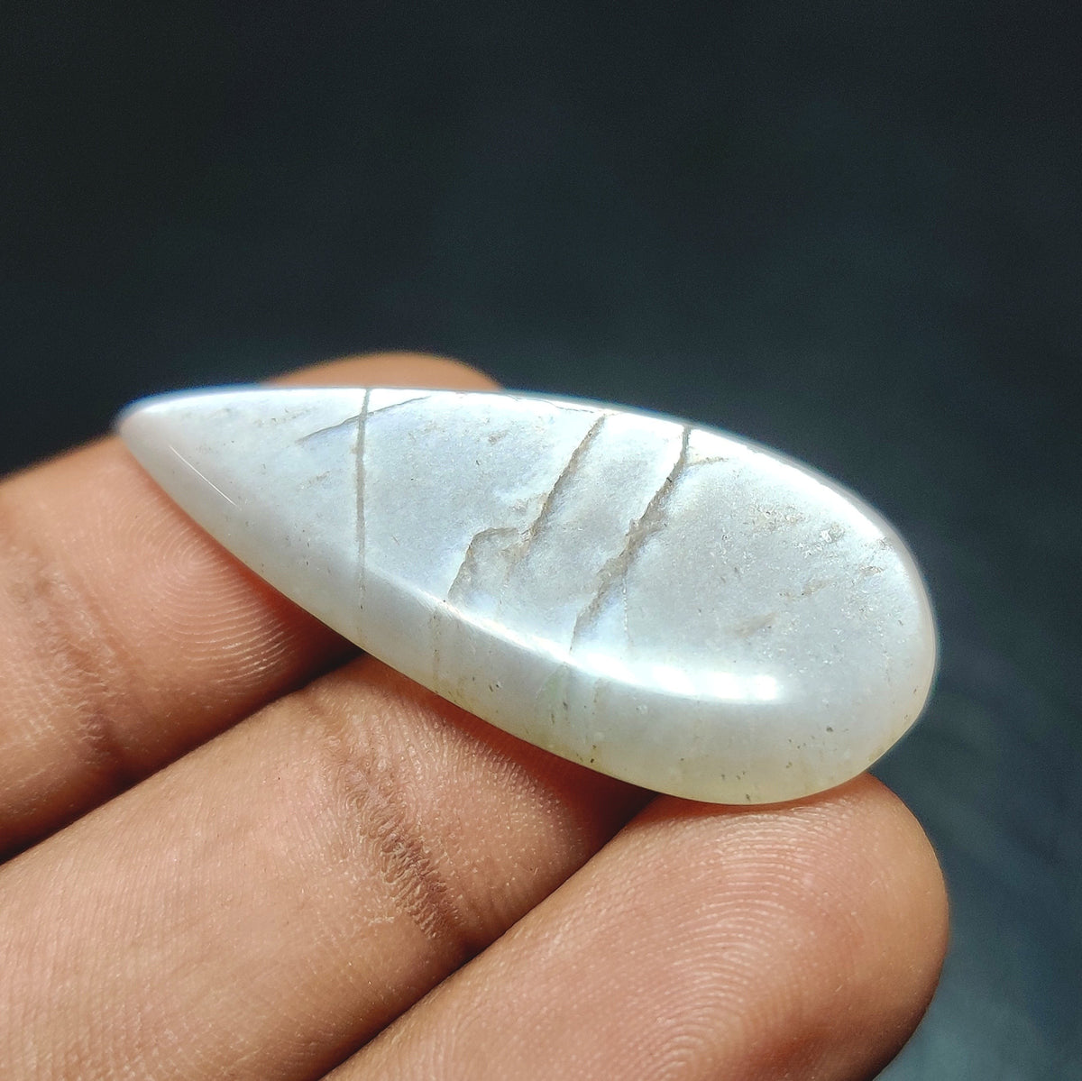 Cabochons4sale pear-shaped translucent grey moonstone cabochon held between fingers against a dark background